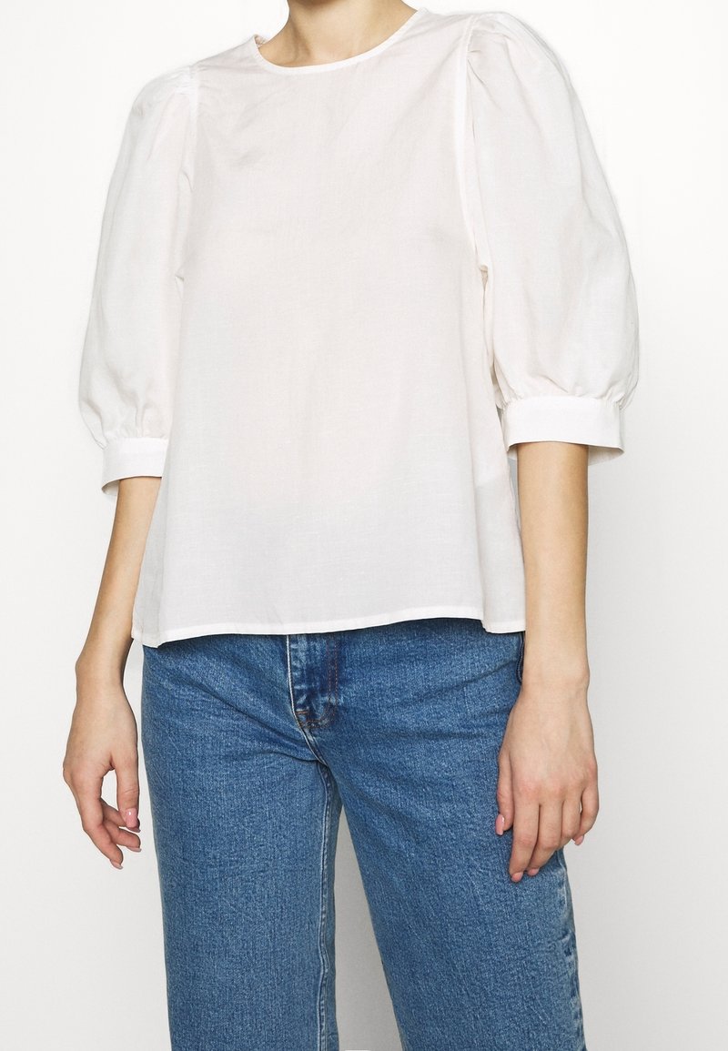 Personne portant un chemisier blanc ample avec des manches trois-quarts bouffantes et un jean en denim bleu, debout devant un fond clair uni.