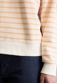 Bluza w paski wykonana z miękkiego materiału, z jasnopomarańczowymi paskami na kremowym tle, wyposażona w białe prążkowane mankiety i pas biodrowy.