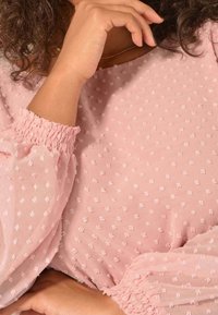 Blusa rosa chiaro con pois a rilievo, maniche elastiche e scollo rotondo. Il tessuto trasparente esalta la delicatezza del design.
