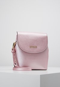 Vaaleanpunainen metallinhohtoinen mini-reppu, jossa on pyöreä läppä, kultaiset metalliosat ja säädettävä olkahihna. Edessä brändin nimi "GUESS kids".