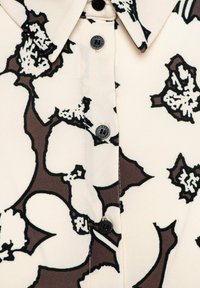 Overhemdblouse met een crèmekleurige achtergrond, versierd met grote zwarte bloemenpatronen en donkerbruine accenten. Zwarte knopen accentueren de sluiting.