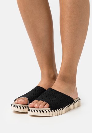 Sandales à enfiler noires perforées avec bout ouvert, dotées d'une semelle ondulée de couleur crème et de coutures noires contrastantes.