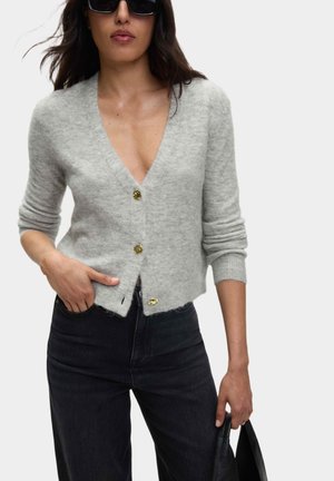 Cardigan in maglia grigia con scollo a V profondo, tre bottoni dorati e polsini a coste. Abbinato a jeans scuri e una borsa nera. Texture liscia.