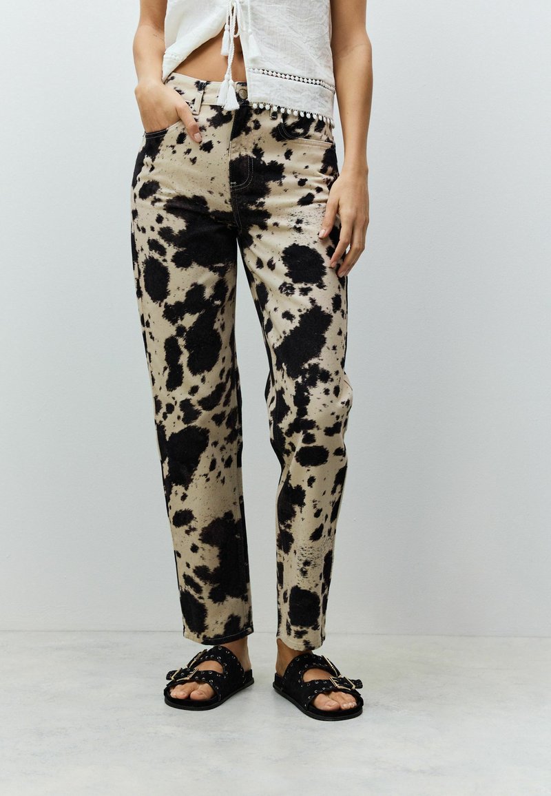 Beige und schwarz gepunktete Jeanshose mit geradem Bein, ausgestattet mit Seitentaschen und schwarzem Nähten, kombiniert mit schwarzen Sandalen.