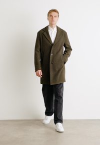 Olivgrön yllekappa med snedskurna slag, enkel knappstängning, sidofickor, i kombination med svarta byxor och vita sneakers mot en neutral bakgrund.