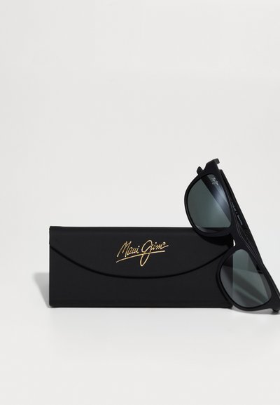 Czarne okulary przeciwsłoneczne Maui Jim z ciemnymi soczewkami, z teksturowanym wzorem ramki, leżące na czarnym etui z złotym logo.
