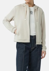 Beige Zip-up-Hoodie mit Kapuze, Kordelzügen und Fronttaschen. Aus glattem Stoff gefertigt, kombiniert mit dunkelblauen Jeans.