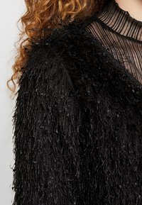 Svart fransad jacka med strukturerad tyg; har en blandning av långa och korta fibrer. Genomskinlig svart krage med horisontella ränder vid halsringningen.