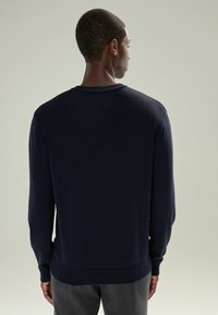 Bărbat �îmbrăcat cu un pulover simplu, lung cu mânecă, de culoare bleumarin închis, și pantaloni gri, stând cu spatele în fața unui fundal simplu, deschis.