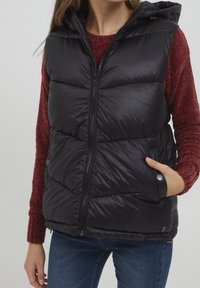 Svart vadderad väst med dragkedja fram, huva och sidofickor; bärs över en röd strukturerad tröja. Har en glänsande yta och quiltat mönster.