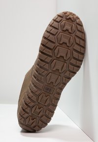 Braune Gummisohle mit einem strukturierten Profil, das hexagonale und keilförmige Formen für Trittfestigkeit aufweist. Verstärkte Ränder und sichtbare Logo-Details.