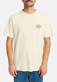 Camiseta de algodón color crema con un pequeño gráfico de un ojo en rosa y azul en el lado izquierdo del pecho. Mangas cortas y ajuste relajado.