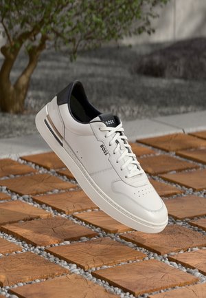Witte en zwarte BOSS sneaker met veters, zwevend boven een patroon van hout en kiezelstenen buitenoppervlak, nabij groen Foliage.