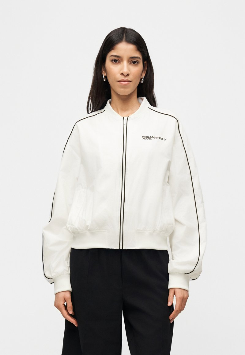 Γυναίκα που φοράει λευκό μπουφάν με φερμουάρ και μαύρες ρίγες, με το λογότυπο "Karl Lagerfeld Jeans", συνδυασμένο με μαύρο παντελόνι, στέκεται μπροστά σε απλό φόντο.