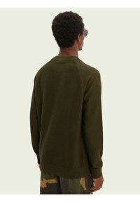 Man met kort krullend haar, gekleed in een olijfgroene trui en camouflagebroek, met de rug naar de camera, tegen een effen lichte achtergrond.