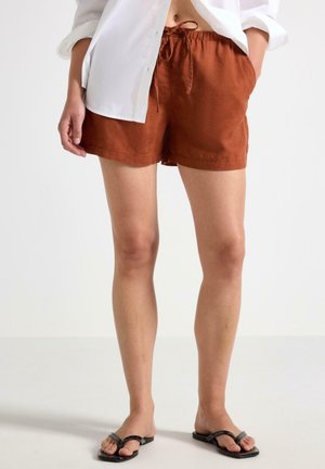 Kvinde iført rustfarvede shorts med snøre, hvid skjorte og sorte klipklappere, står med den ene hånd i lommen på en neutral baggrund.