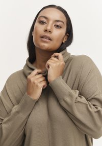 Pull en khaki oversize avec col cheminée, manches côtelées et tissu texturé, les mains tenant l'encolure, sur un fond uni.