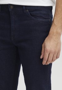 Jeans de mezclilla azul oscuro con un ajuste slim. Cuentan con un bolsillo frontal y un cierre de botón, resaltados por sutiles detalles de costura.