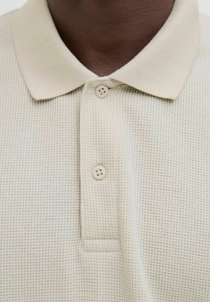 Béžová polo košela s textúrovanou tkaninou, obsahujúca trojbuttonsko a mäkký golier. Materiál má jemný mriežkový vzor.