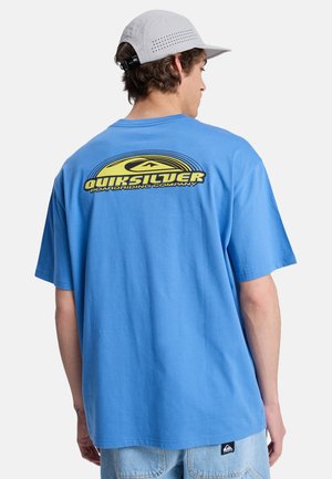 Mladi moški v modri majici Quiksilver z rumenim logotipom na hrbtu in svetlo sivo kapo, obrnjeni stran, na belem ozadju.