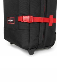 Černý cestovní batoh na kolečkách vyrobený z odolné tkaniny, s červeným popruhem na přezku a logem Eastpak, zaobleným dnem a ochrannými nožičkami.