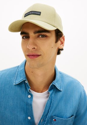 Ung mand iført en beige Tommy Hilfiger kasket, blå denimskjorte med logo på lommen og hvid T-shirt mod en ensartet hvid baggrund.