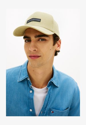 Νεαρός άνδρας με μπεζ καπέλο Tommy Hilfiger, μπλε τζιν πουκάμισο με λογότυπο στην τσέπη και λευκό T-shirt, σε απλό λευκό φόντο.