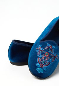 Zapatillas de terciopelo azul con bordado floral en rojo, verde y azul. Diseño de punta redondeada con una suela plana negra. Textura suave.