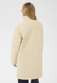 Long manteau matelassé beige clair, avec un col montant, des épaules tombantes et un motif texturé avec des surpiqûres ondulées.
