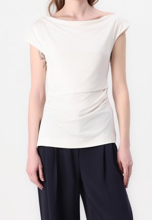 Vrouw draagt een wit gedrapeerd mouwloos topje en zwarte broek met wijde pijpen, staand tegen een effen witte achtergrond.