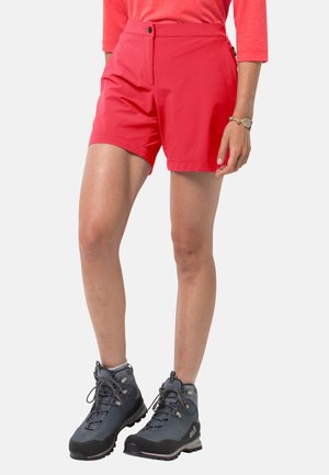 Frau trägt rote Shorts bis zur Mitte des Oberschenkels, korallenfarbenes langärmeliges Oberteil, graue Wanderschuhe und Armband, steht vor einem einfarbigen Hintergrund.