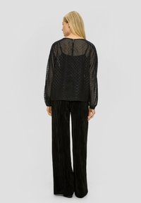 Schwarzes, durchsichtiges Oberteil mit dezenten Punkten, runder Ausschnitt und langen Ärmeln. Kombiniert mit schwarzen, plissierten Hosen. Einfacher Schlitz im Rücken.