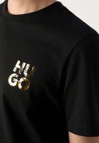 T-shirt en coton noir avec un logo "HUGO" doré à texture fissurée sur la poitrine gauche. Manches courtes et col rond.