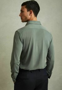 Cămașă cu mânecă lungă dintr-un material moale, de un verde estompat, cu o textură netedă, având un guler pliabil și manșete cu nasturi, asortată cu pantaloni închiși la culoare.