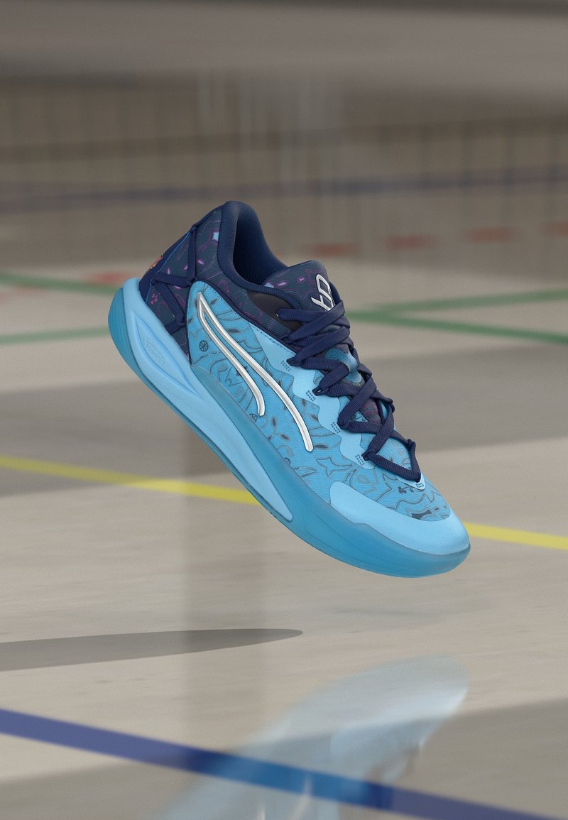 Μπλε και ναυτικό αθλητικό παπούτσι με σχεδιασμένο επάνω μέρος και λευκή ρίγα, στον αέρα σε εσωτερικό αθλητικό γήπεδο με έγχρωμες γραμμές.