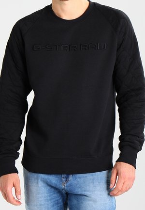 Zwarte sweatshirt met getextureerde gewatteerde mouwen, gevormd logo op de borst, ronde hals en geribde manchetten, gecombineerd met lichtblauwe jeans.