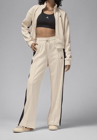 Conjunto de chaqueta y pantalones de cremallera beige con rayas laterales negras. Sujetador deportivo negro debajo de la chaqueta. Presenta el logo de Jordan en la chaqueta y en los pantalones.