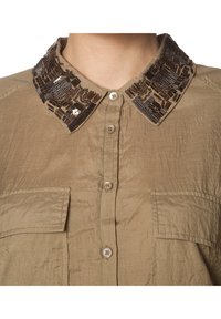 Camisa clara de botones con cuello texturizado y con lentejuelas de patrones intrincados, bolsillo frontal y un acabado de tela ligera.