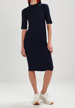 Robe midi bleu marine côtelée avec manches courtes et col haut. Portée avec des chaussures blanches à semelle marron clair.