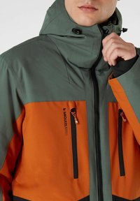 Herenjack met een waterdicht ontwerp, gekleurd in grijs en oranje, met een verstelbare capuchon, ritszakken en een gestructureerde stof.