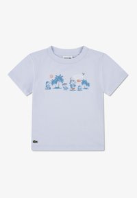 Ανοιχτό μπλε βαμβακερό μπλουζάκι με κοντά μανίκια. Στο μπροστινό μέρος υπάρχει γραφικό με φοίνικες, ήλιο και παιχνιδιάρικους χαρακτήρες. Λογότυπο Lacoste στο τελείωμα.