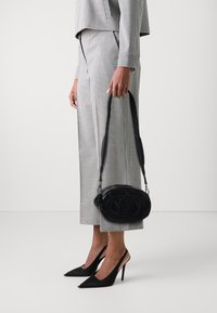 Zwart leren ovale handtas met reliëflogo, crossbody gedragen. Het model draagt een zwart-wit houndstooth pak en spitse zwarte hakken.
