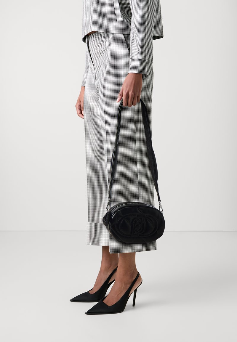 Zwart leren ovale handtas met reliëflogo, crossbody gedragen. Het model draagt een zwart-wit houndstooth pak en spitse zwarte hakken.
