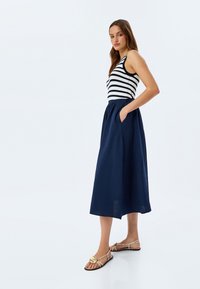 Top senza maniche a righe abbinato a una gonna midi blu navy con tasche. Modella indossa sandali con dettagli dorati su uno sfondo bianco.