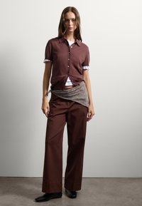 Cardigan corto marrone con maniche corte e bottoni, indossato sopra una maglietta bianca. Abbinato a pantaloni marroni a vita alta e a una sciarpa decorata.