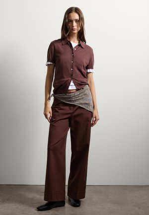 Bruine korte mouwen cardigan met knopen, over een wit T-shirt. Gecombineerd met hoge, bruine broeken en een gebloemde sjaal.