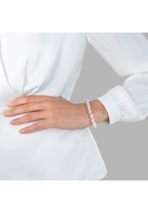 Pulsera de perlas que presenta perlas redondas y lisas de diferentes tamaños, exhibida en una mano reposando sobre una blusa blanca de tela texturizada.