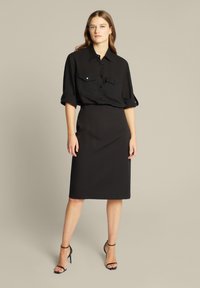 Schwarze Hemdjacke mit zwei Brusttaschen, hochgeschobenen Ärmeln, kombiniert mit einem schwarzen Bleistiftrock und schwarzen, hochhackigen Sandalen mit Riemchen.