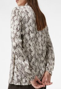 Woman with long brown hair wearing a beige and brown snake print long-sleeve blouse, hands clasped behind back, standing sideways.