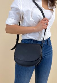 Borsa a tracolla in pelle nera con pattina arrotondata, texture liscia e tracolla regolabile, indossata da una persona su uno sfondo neutro.